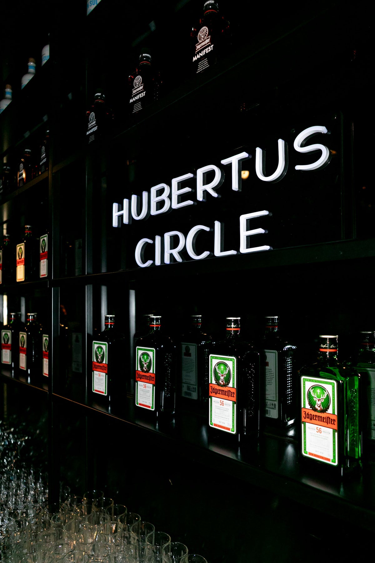 A row of Jägermeister bottles displayed on dark shelves under a glowing white HUBERTUS CIRCLE sign.