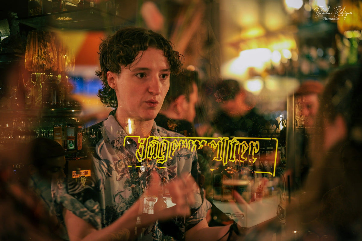A bartender in a floral shirt works behind a glass partition reflecting a neon Jägermeister sign and the warm lights of a busy bar.