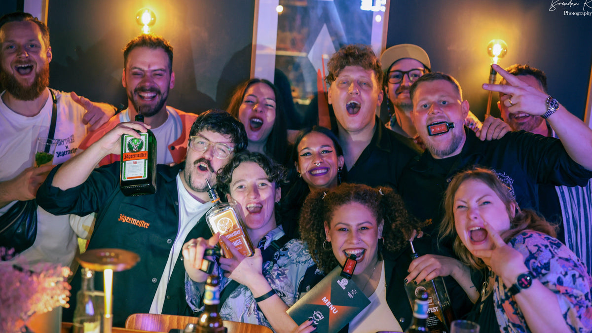 A group of nine people posing together in a dimly lit bar or club environment, holding up bottles of alcohol and smiling, laughing, and making funny faces. The scene is illuminated by warm, glowing Edison bulbs hanging in the background.