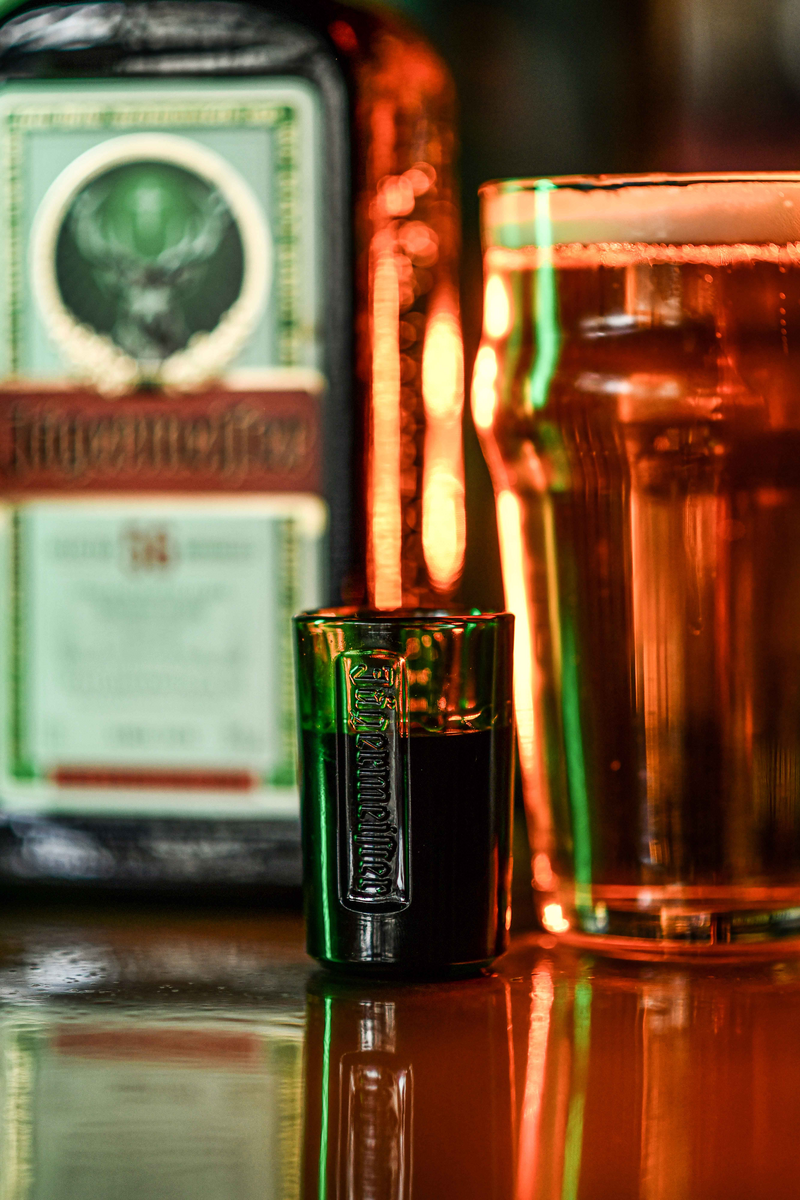 A Jägermeister shot glass with embossed banding in the foreground, with an beer glass in the back and a blurred Jägermeister bottle in the background on a reflective bar surface.