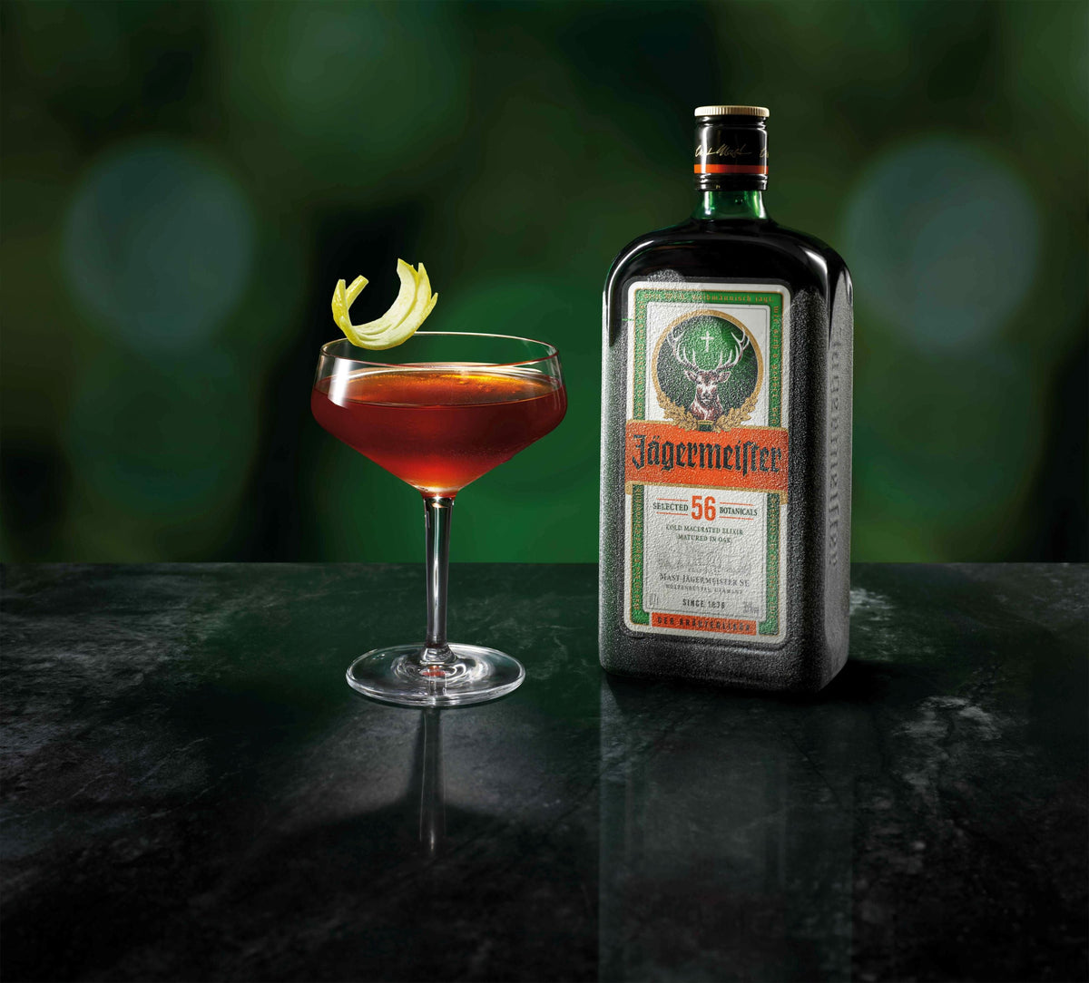 A bottle of ice cold Jägermeister next to a coupe glass filled with an amber-red cocktail and a lemon twist garnish, set on a dark reflective surface against a blurred green background.