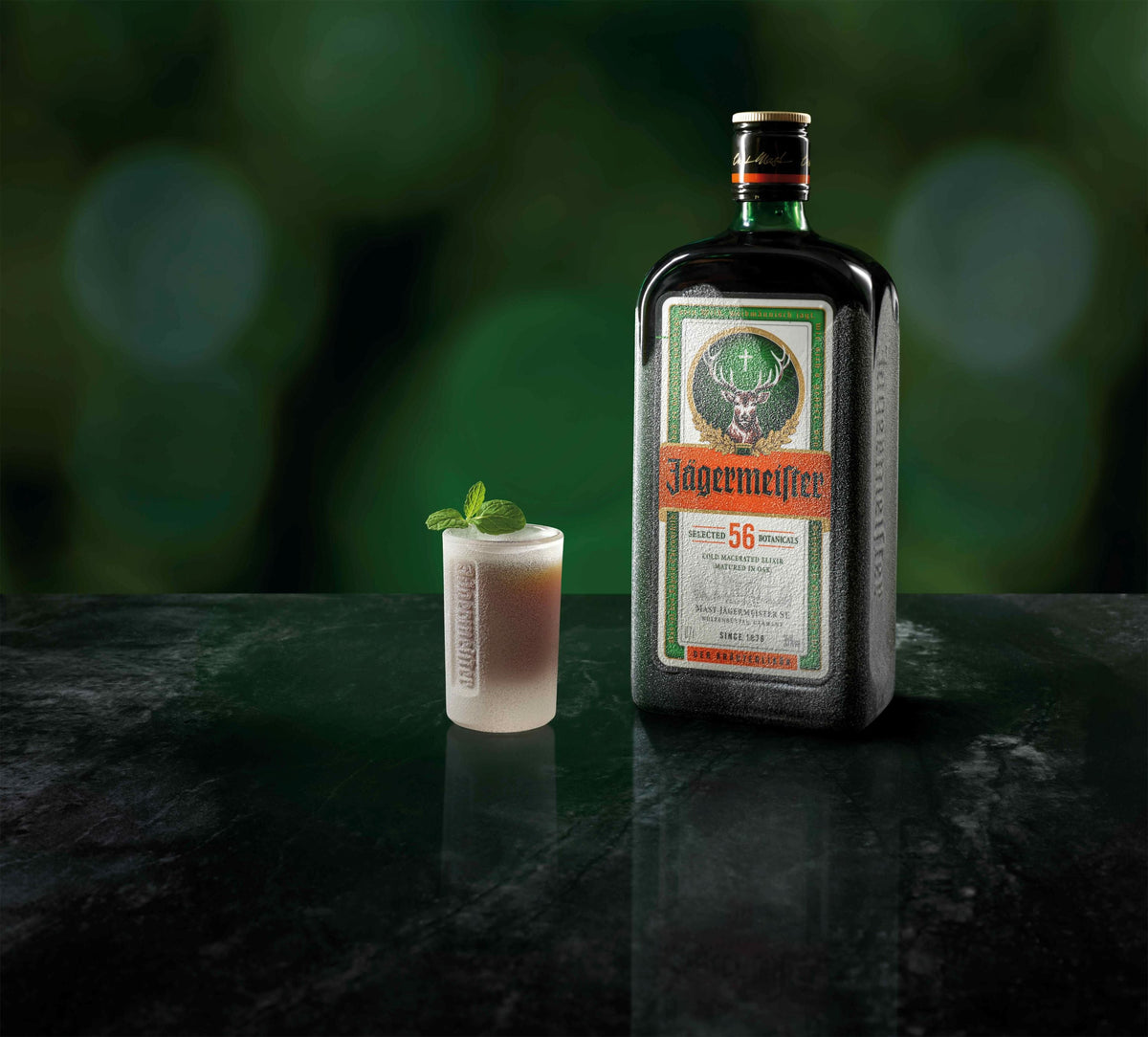 A bottle of Jägermeister stands on a dark reflective surface beside a frosted shot glass and garnished with a mint leaf, against a blurred green background.