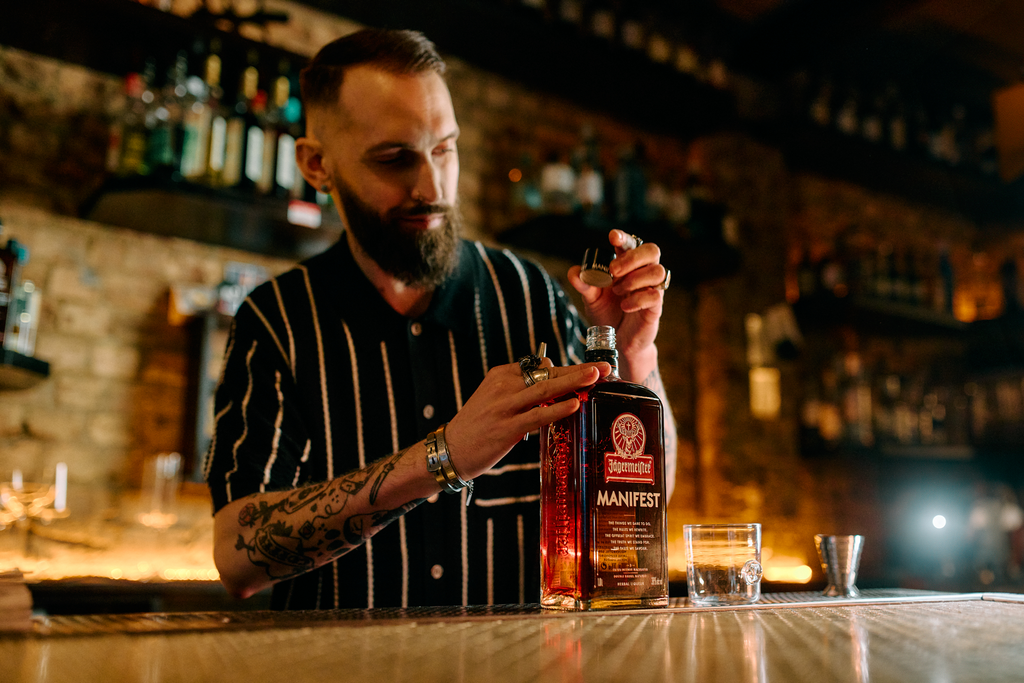 A bearded man in a black and white striped shirt stands behind a bar counter, holding a bottle of Manifest next to a small glass and a metal shot glass, with a warm, dimly lit bar background.