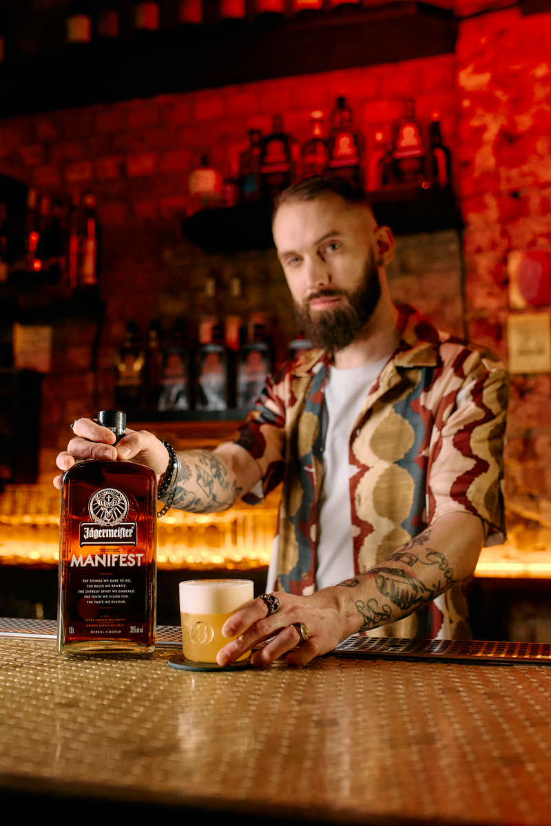 A man with a beard and tattoos stands behind a bar holding a bottle of Manifest next to a glass with a cocktail, with a warm, red-lit bar background.