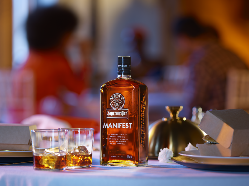 A bottle of Fengermae Manifest whiskey stands on a table in a restaurant setting, with two glasses of the amber liquid, a blurred background showing people dining, and a gold cloche.