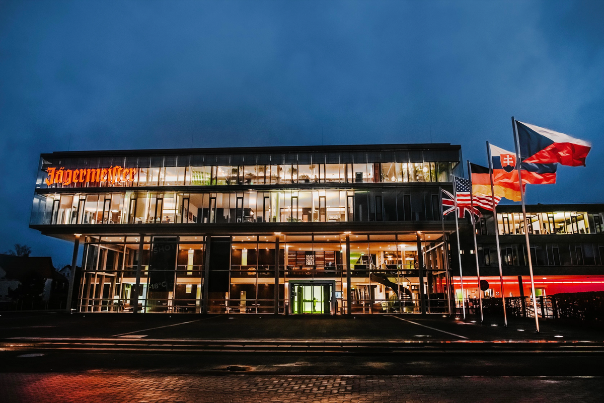 The exterior of the Jägermeister headquarters building at dusk, featuring a modern glass facade with warm interior lighting, a prominent orange Jägermeister logo, and several flags on poles to the right against a deep blue evening sky.