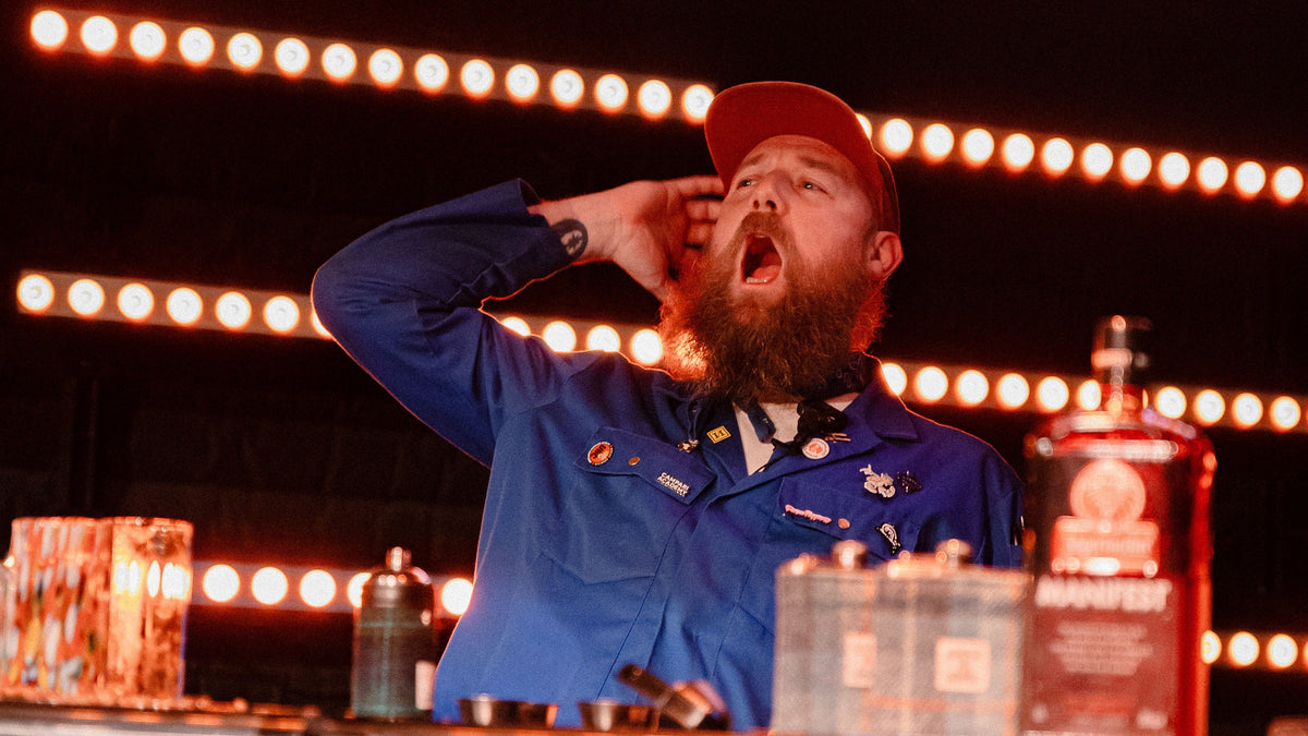 A bearded man in a blue work shirt and red cap poses dramatically behind a bar counter with bottles and glowing light bars in the background.
