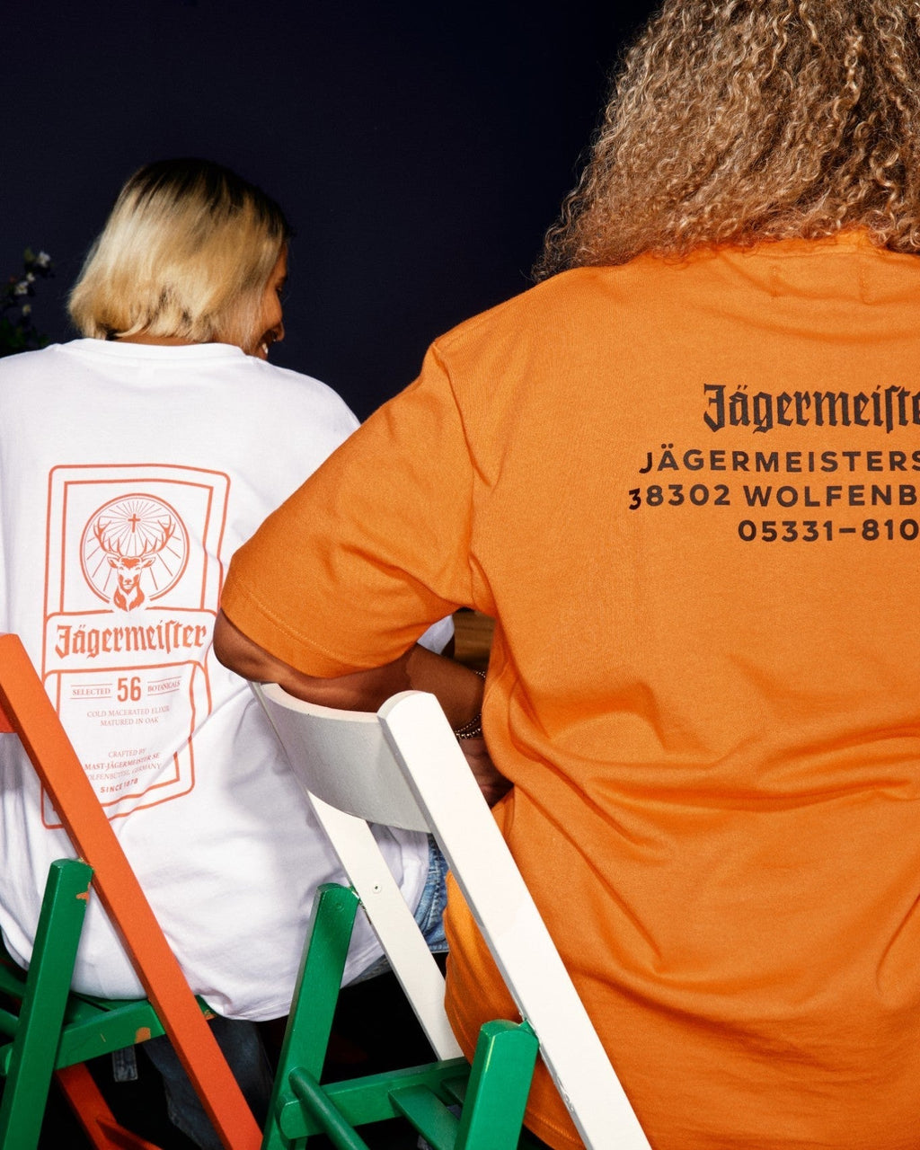 Két ember ül zöld és fehér székeken, hátulról láthatók, Jägermeister márkázott pólóban. Az egyik póló fehér, narancssárga címkenyomattal, a másik élénk narancssárga, rajta a Jägermeister központjának címe feketével.