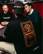 Négy ember gyűlt össze egy asztal körül egy ételstandnál, a középpontban egy fekete Jägermeister pólót viselő személy áll, akinek a hátán nagy narancssárga grafika van, khaki nadrágot és fekete sapkát visel. 