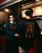 Két férfi áll egy piros food truck előtt éjszaka. Az egyik férfi a kamerába néz, sötétzöld Jägermeister hosszú ujjú felsőt és fekete nadrágot visel. A másikon a Jägermeister Racing Cap és a fekete Jägermeister címkés póló van.