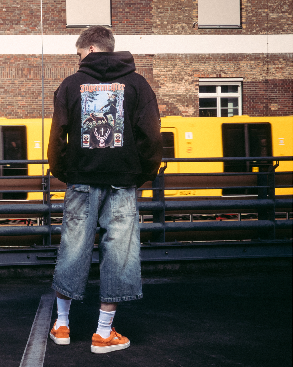 Egy modell streetwear ruházatban, többek között fekete kapucnis pulóverben, Jägermeister tradition grafikus hátoldali nyomattal, egy sötét burkolt felületen áll egy téglaház és egy sárga vonat előtt.
