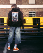 Egy modell streetwear ruházatban, többek között fekete kapucnis pulóverben, Jägermeister tradition grafikus hátoldali nyomattal, egy sötét burkolt felületen áll egy téglaház és egy sárga vonat előtt.