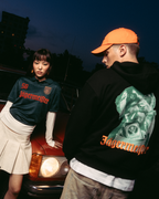 Two models posing in front of a vintage red car at dusk. One model wears a dark green Jägermeister jersey with a white pleated skirt, and the other wears a black Jägermeister Waldi hoodie and an orange Jägermeister Dad cap.
