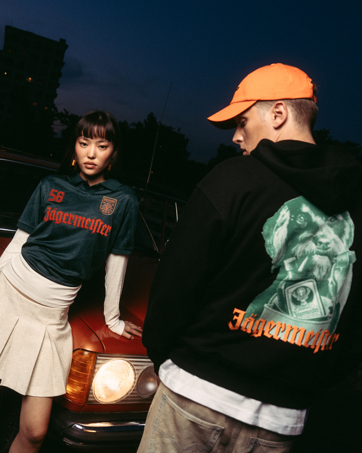 Két modell pózol egy vintage piros autó előtt alkonyatkor. Az egyik modell sötétzöld Jägermeister mezt visel fehér rakott szoknyával, a másik fekete Jägermeister Waldi kapucnis pulóvert és narancssárga Jägermeister Dad Capet.