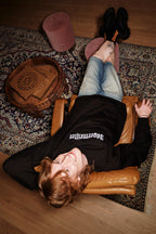 Person lying on a leather chair with feet up on a rug, wearing a black Jägermeister crewneck.
