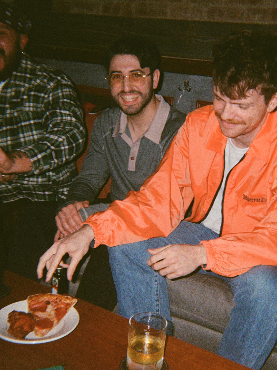 Két férfi ül együtt, az egyiken narancssárga Jägermeister kabát van, az asztalon egy pizzaszelet és egy pohár látható.