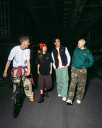 Négy ember sétál és biciklizik egy sötét utcán éjszaka, egy magasvasút alatt, különböző színű Jägermeister streetwear ruházatban.
