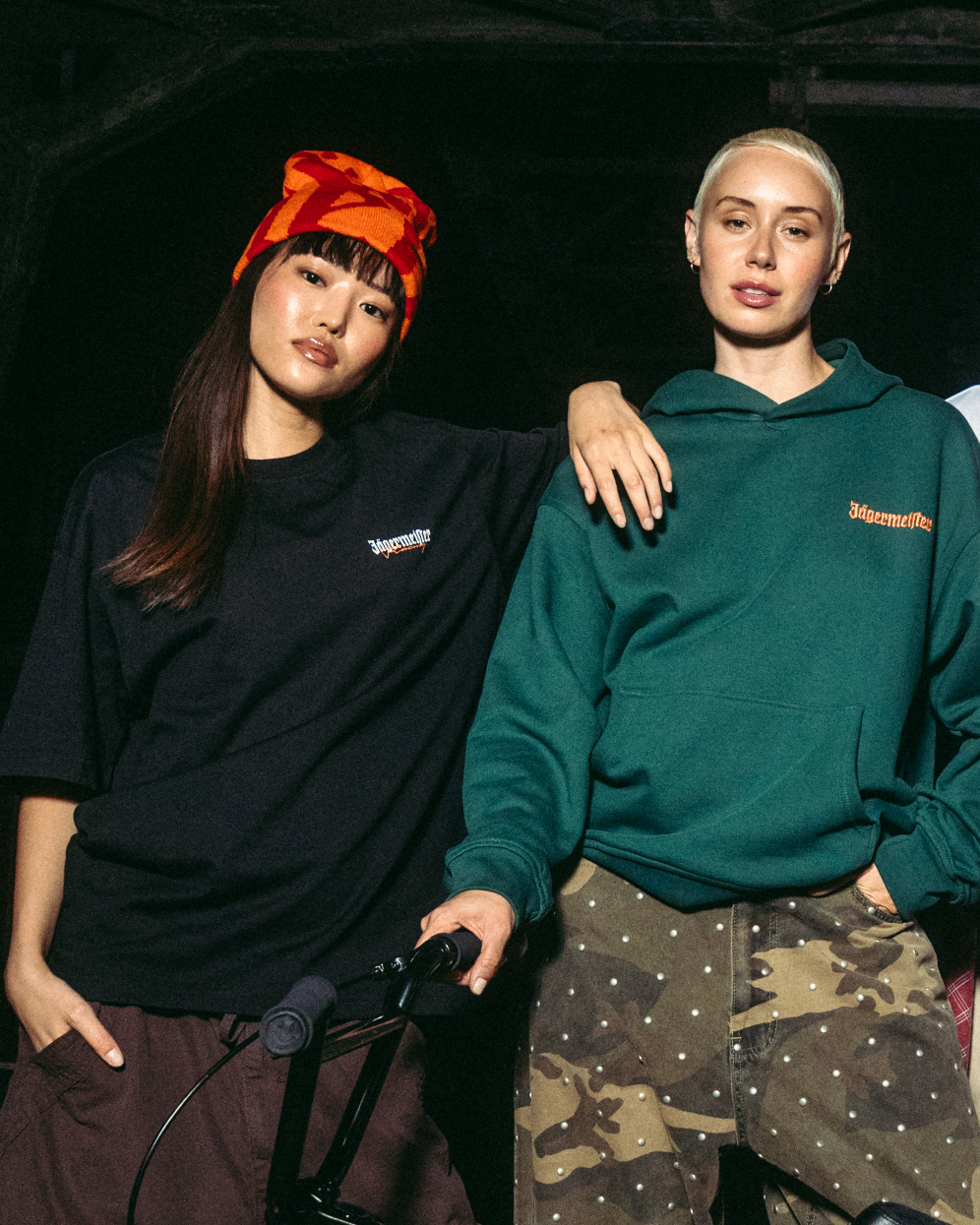 Két fiatal nő pózol egy fekete BMX biciklivel egy sötét ipari híd előtt éjszaka. Jägermeister streetwear ruházatot viselnek, többek között zöld kapucnis pulóvert, fehér pólót, fekete mellényt és cargo nadrágot.