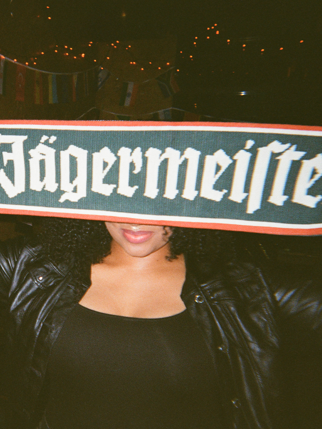 Személy Jägermeister sállal az arca előtt, sötét háttér előtt.
