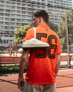 Egy személy szabadtéri sportpályán áll, élénk narancssárga Jägermeister mezt visel, hátul nagy, nyomtatott 56-os számmal. Az egyik vállán egy pár fehér Jägermeister sneaker lóg, a háttérben drótkerítés és lakóházak láthatók.