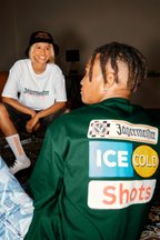 Two people sitting indoors, one wearing a white shirt with Jägermeister branding, the other with a green jacket featuring Jägermeister.