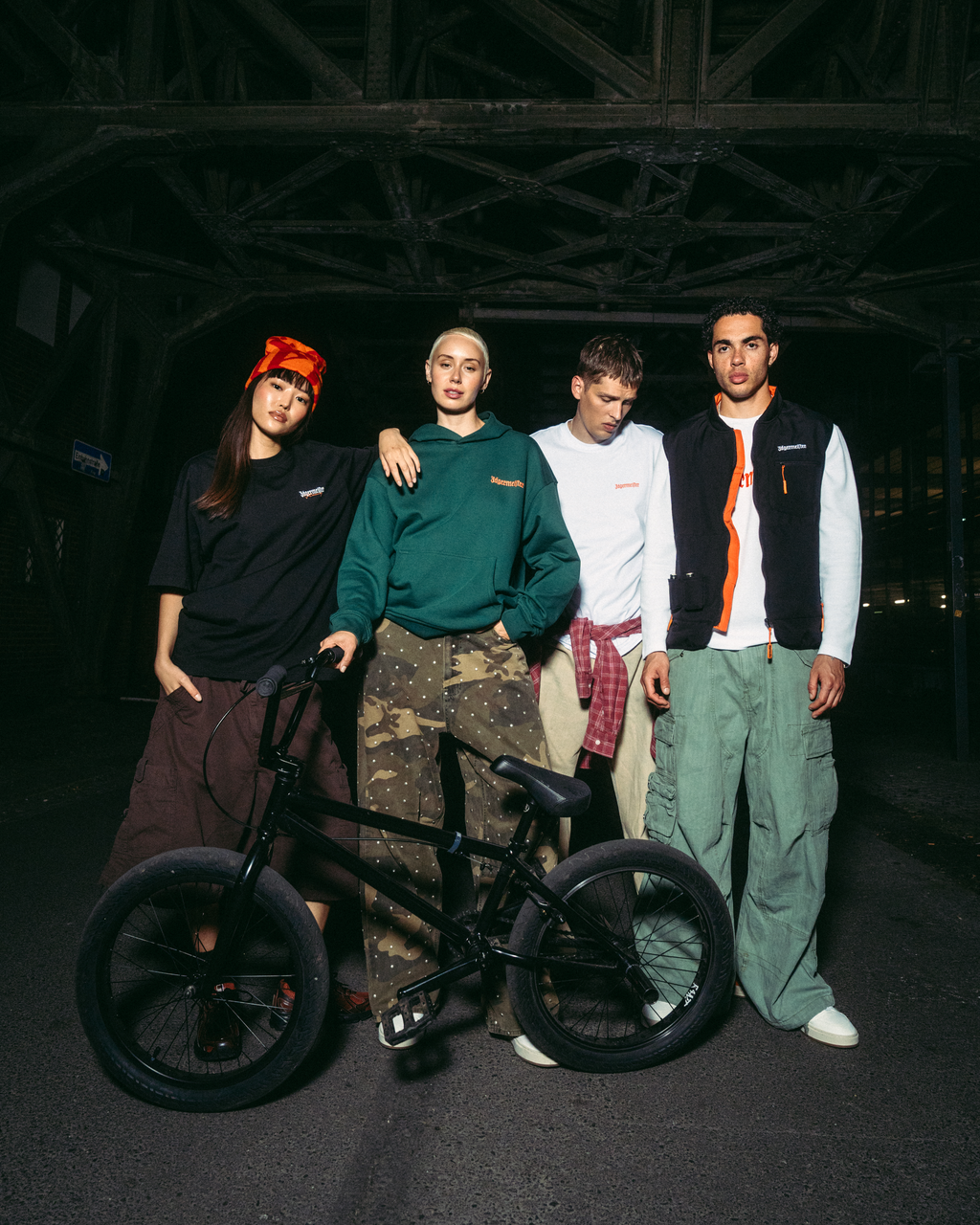 Négy fiatal férfi pózol egy fekete BMX biciklivel egy sötét, ipari hídnál éjszaka. Jägermeister utcai viseletet viselnek, többek között zöld kapucnis pulóvert, fehér pólót, fekete mellényt és cargo nadrágot.