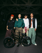 Négy fiatal férfi pózol egy fekete BMX biciklivel egy sötét, ipari hídnál éjszaka. Jägermeister utcai viseletet viselnek, többek között zöld kapucnis pulóvert, fehér pólót, fekete mellényt és cargo nadrágot.