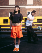 Four models wearing Jägermeister streetwear apparel, including orange shorts, a black crop top, a white t-shirt, and a black hoodie with a graphic back print, standing on a dark paved surface in front of a brick building and a yellow train.