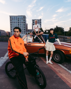 Three models in Jägermeister streetwear outfits posing with a vintage orange car and a black BMX bike on an urban rooftop at sunset. The outfits include a bright orange Jägermeister zipper jacket, black track pants with red stripes, a white Jägermeister jersey, a green Jägermeister jersey with orange branding and a white pleated skirt.