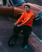 A group of four people in an urban outdoor setting with a rusted orange car, modern high-rise buildings, and a bridge in the background. One person in a bright orange Jägermeister zipper sits on a bicycle in the foreground.