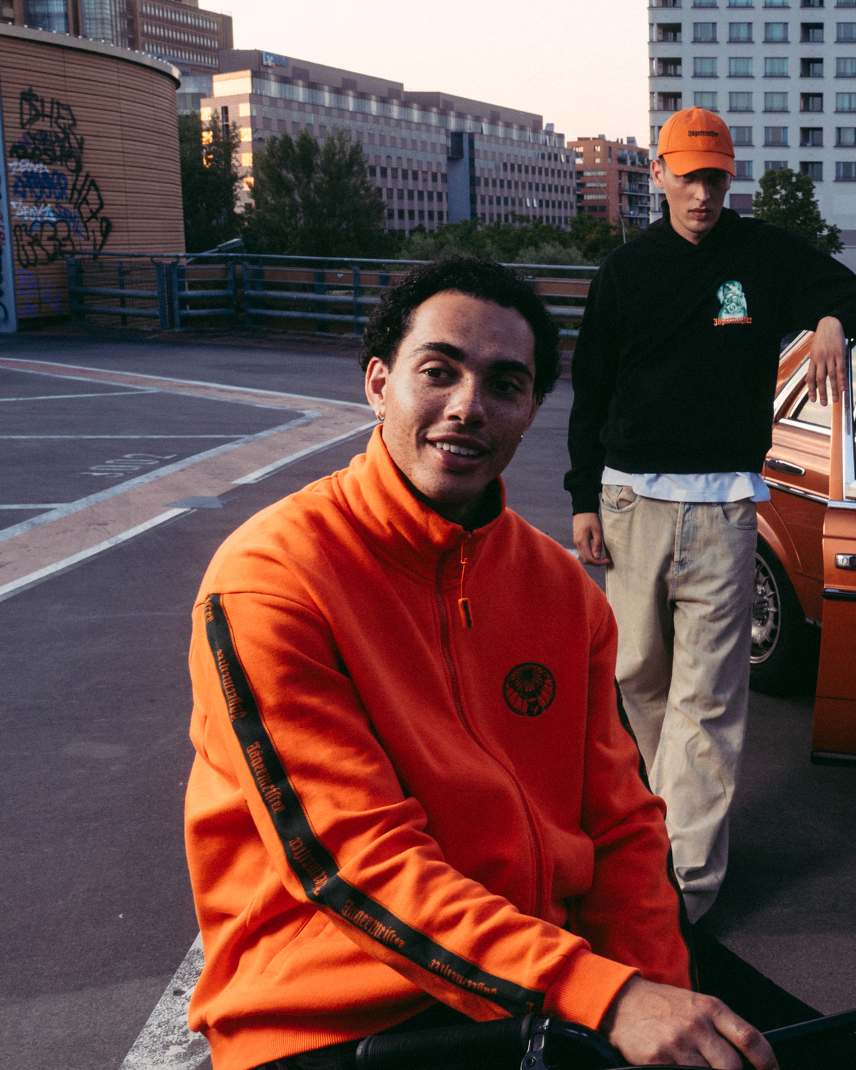 Négy modell pózol egy városi tetőn egy vintage narancssárga autóval, különféle Jägermeister streetwear szettekben, köztük élénk narancssárga Jägermeister cipzáras felsőben, fehér Jägermeister grafikus pólóban Waldival, sötétzöld Jägermeister mezben és fehér rakott szoknyában.