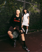 Two models posing in front of a chain-link fence. The model on the left wears a black Jägermeister long-sleeve, brown shorts, and black socks.