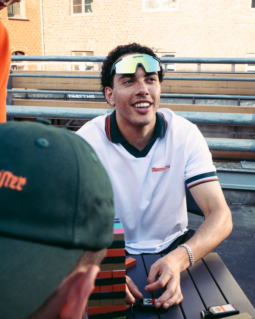 Egy férfi fehér és zöld Jägermeister pólóban mosolyog, miközben egy asztalnál ül a szabadban; az előtérben egy zöld Jägermeier sapkás másik személy látható, a háttérben pedig egy téglaház áll.