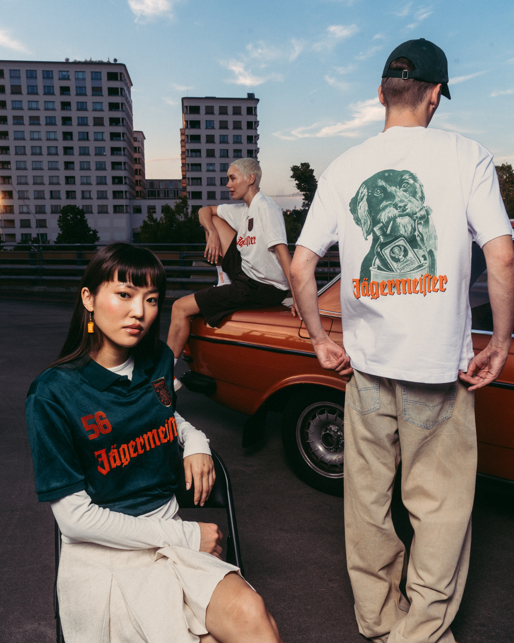 Three models wearing Jägermeister branded apparel in an outdoor urban setting with a vintage orange car and apartment buildings in the background. One model is seated in the foreground wearing a dark green Jägermeister jersey, another stands with his back to the camera in a white graphic T-shirt and beige pants, and a third model sits on the car in a white T-shirt.