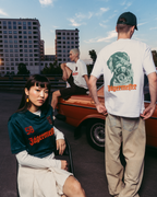 Three models wearing Jägermeister branded apparel in an outdoor urban setting with a vintage orange car and apartment buildings in the background. One model is seated in the foreground wearing a dark green Jägermeister jersey, another stands with his back to the camera in a white graphic T-shirt and beige pants, and a third model sits on the car in a white T-shirt.