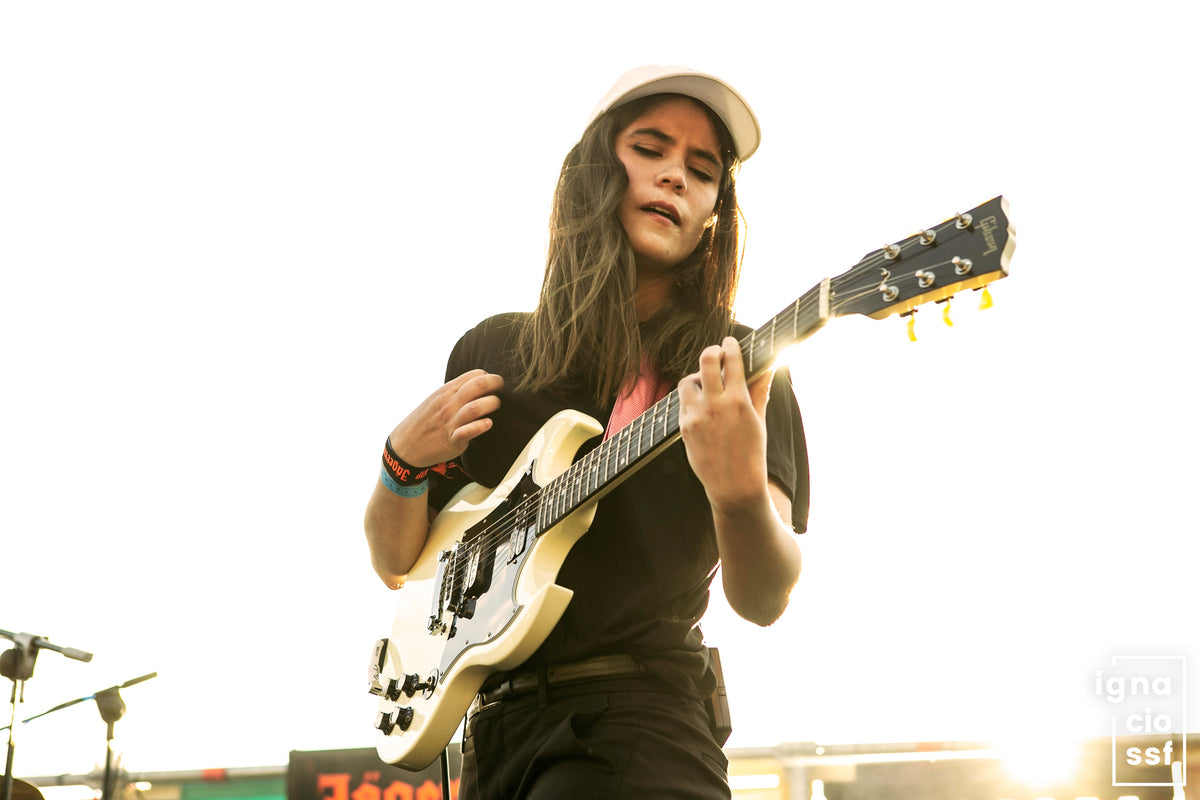 A woman playing an electric guitar on stage, wearing a white cap, black t-shirt, and black pants against a bright, overexposed outdoor background.