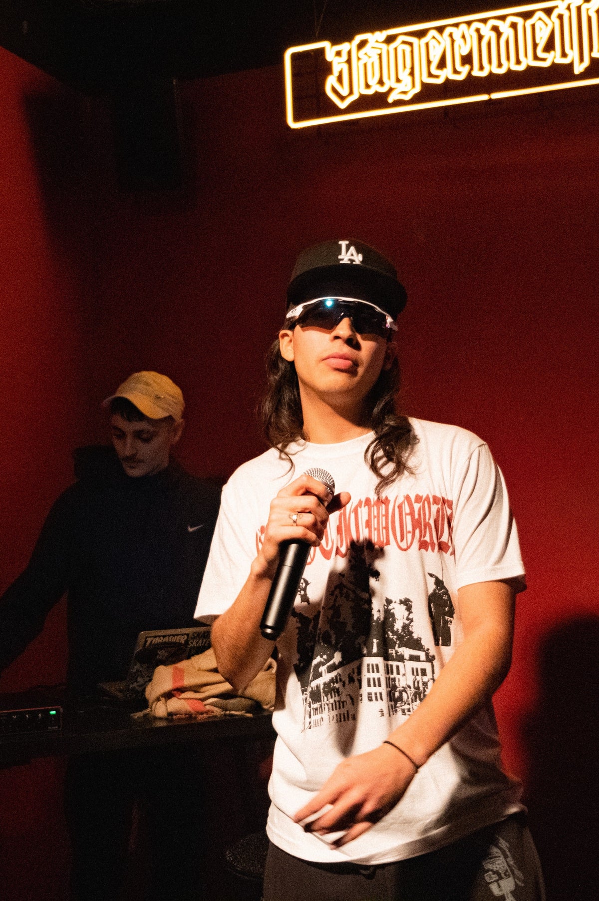 Person holding a microphone and performing in front of a red backdrop, wearing sunglasses and a black cap, with a glowing Jägermeister sign above and another person standing near DJ equipment in the background.