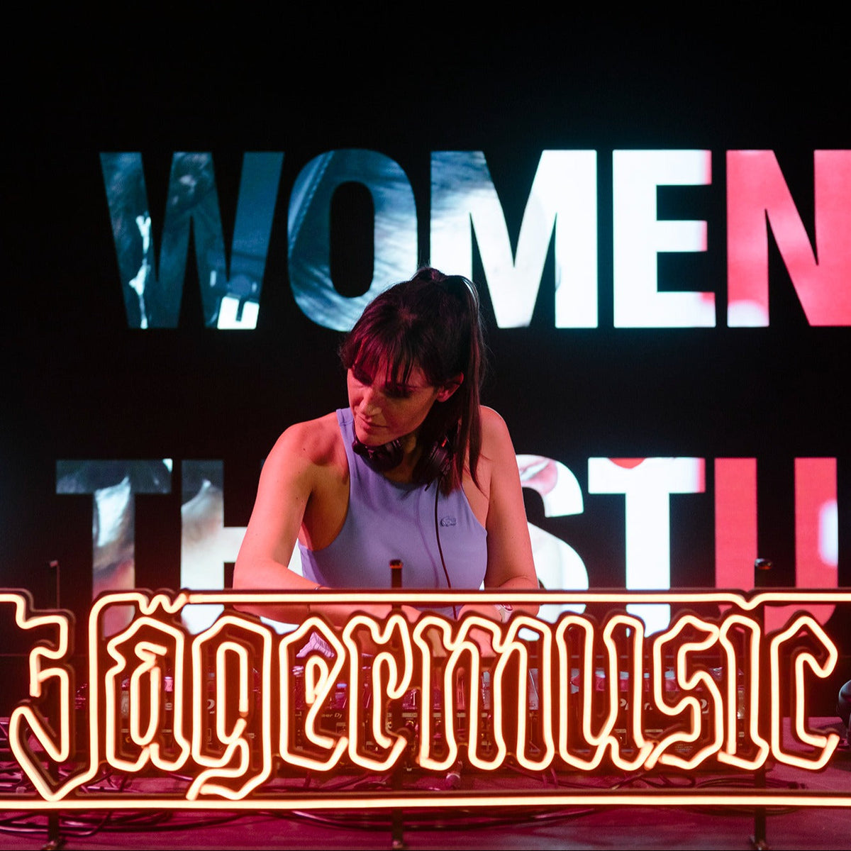 Person wearing headphones standing behind a DJ booth, partially framed by a glowing Jägermeister neon sign, with large illuminated text in the background at an event or stage setting.