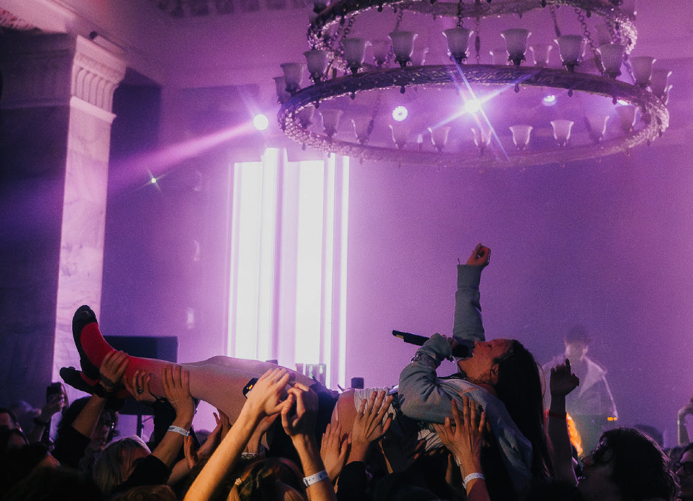 A concert scene with a performer being crowd-sold on stage under a large chandelier, lit by purple stage lights and bright white vertical lights in the background.