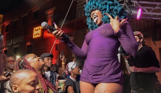 A performer in a purple long-sleeve mini dress with a bright blue afro wig holds a microphone on stage, surrounded by a crowd at an outdoor night event.