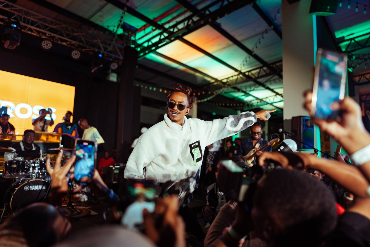 A man in a white hoodie and sunglasses stands on a stage in a crowded event space, pointing toward the audience. A large yellow screen behind him shows the partial brand name CROSS, and multiple people in the foreground hold up smartphones to record the moment.