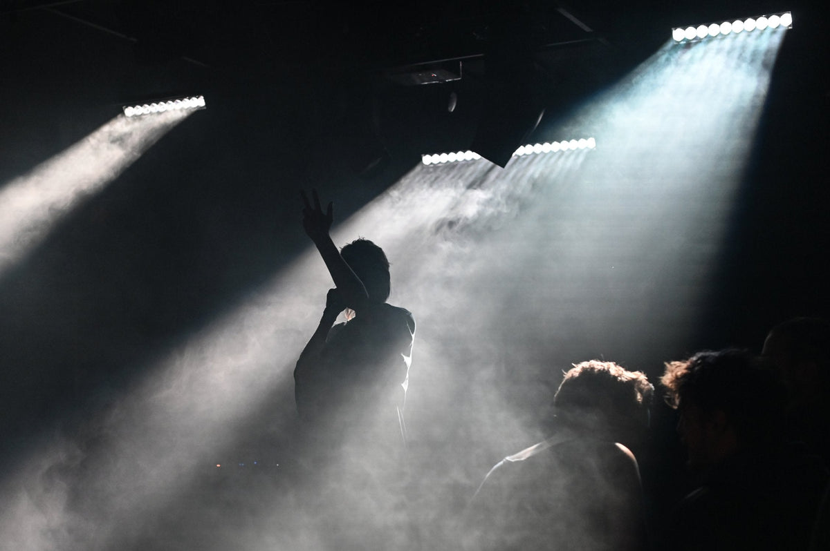 Silhouette of a performer on stage with one arm raised, standing in bright white spotlight beams and surrounded by atmospheric concert fog, with audience figures visible in the foreground.