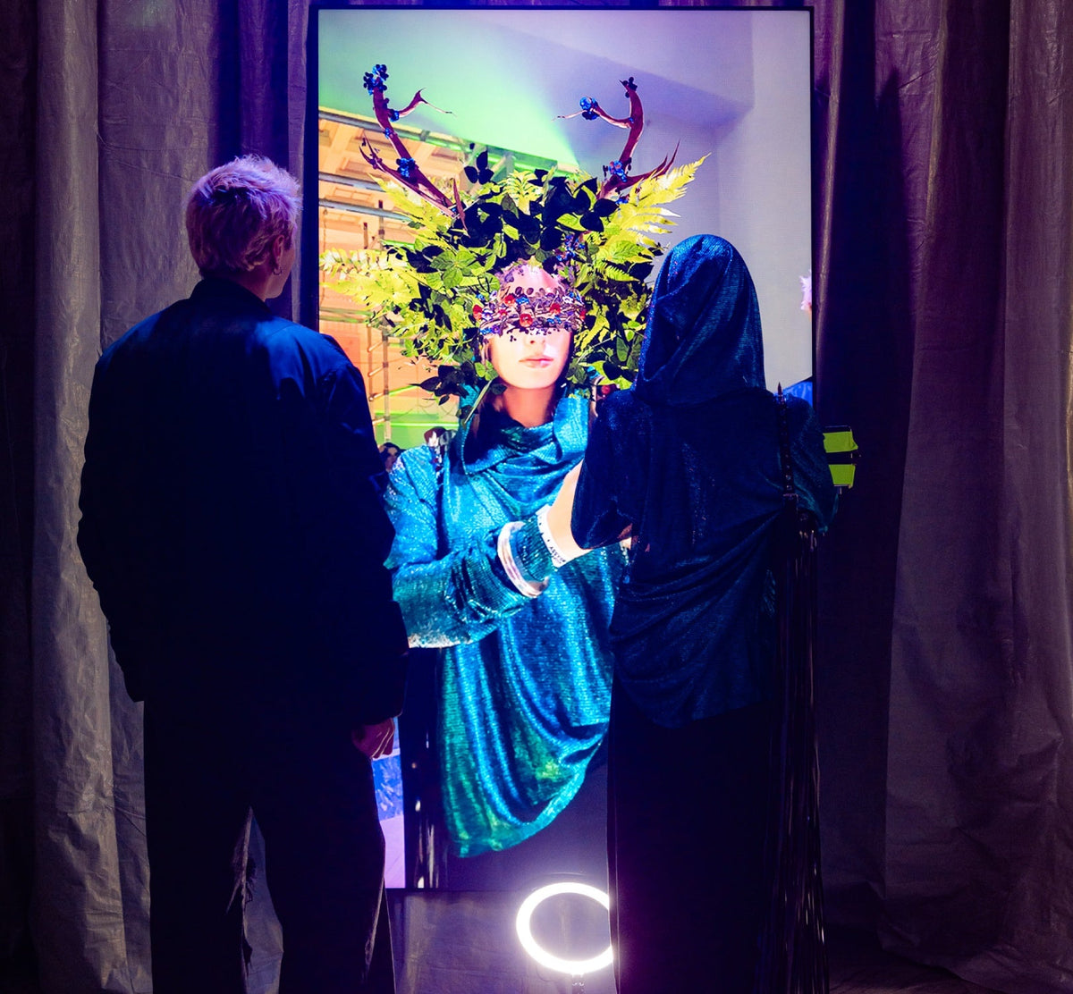 A person in a shimmering teal velvet costume with a decorative mask stands in front of a large mirror, flanked by two people in dark clothing, with curtains framing the scene.