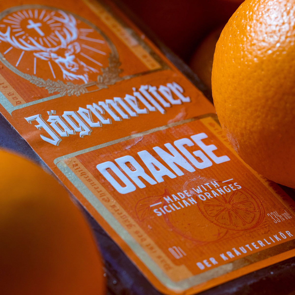 A close-up of a Jägermeistee Orange liqueur bottle label, showing the brand name in gothic lettering and 'ORANGE' in large white text, with fresh orange slices and whole oranges around it.