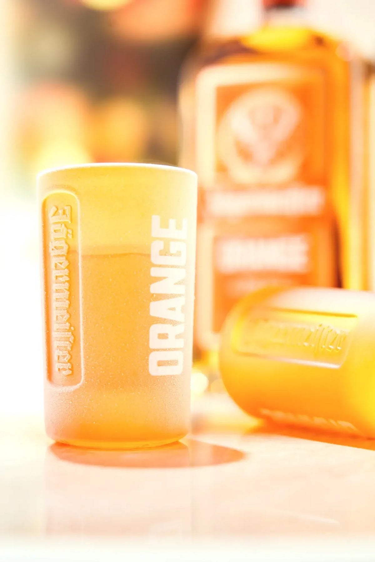 An orange-tinted shot glass with 'ORANGE' printed vertically on the front, shown alongside a Jägermeister Orange bottle in the background.