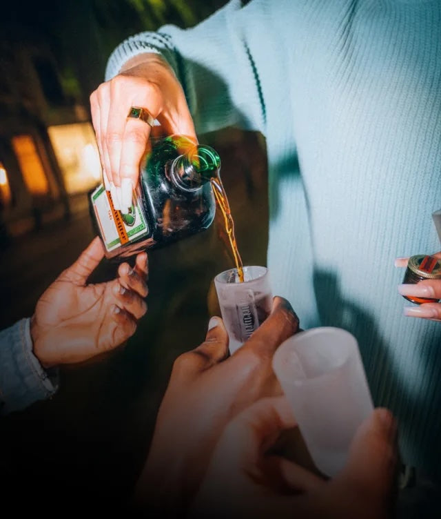 A close-up lifestyle shot of a person pouring Jägermeister into a small frosted cup, with another hand holding a frosted tumbler and a blurred real-world background.