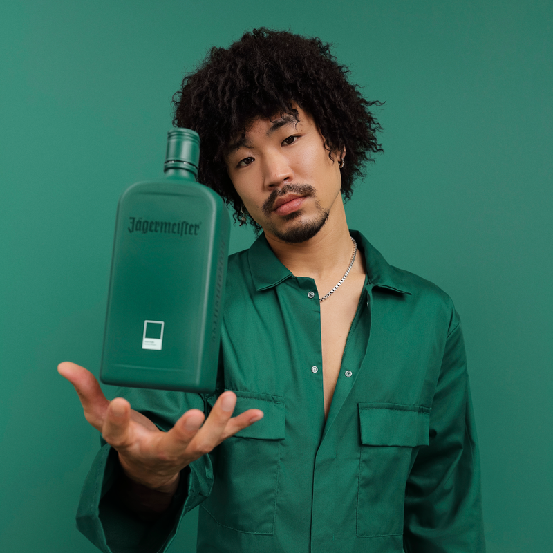 A man with curly dark hair wearing a green button-up shirt holds up a green Jägermeister bottle against a solid green studio background.