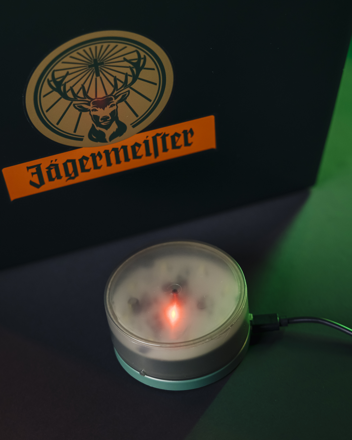 Jägermeister logó sötét háttéren, mellette egy kis, kerek, fényt kibocsátó eszközzel.