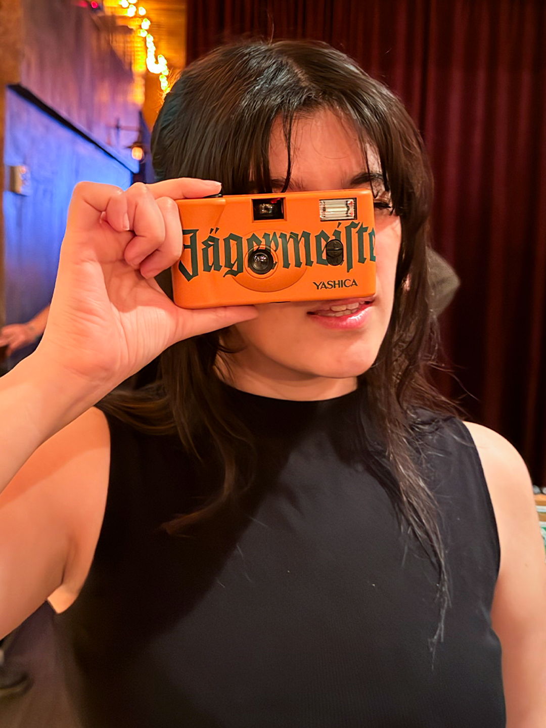 Egy személy narancssárga, „Jägermeister” márkajelzésű fényképezőgépet tart az arca előtt.