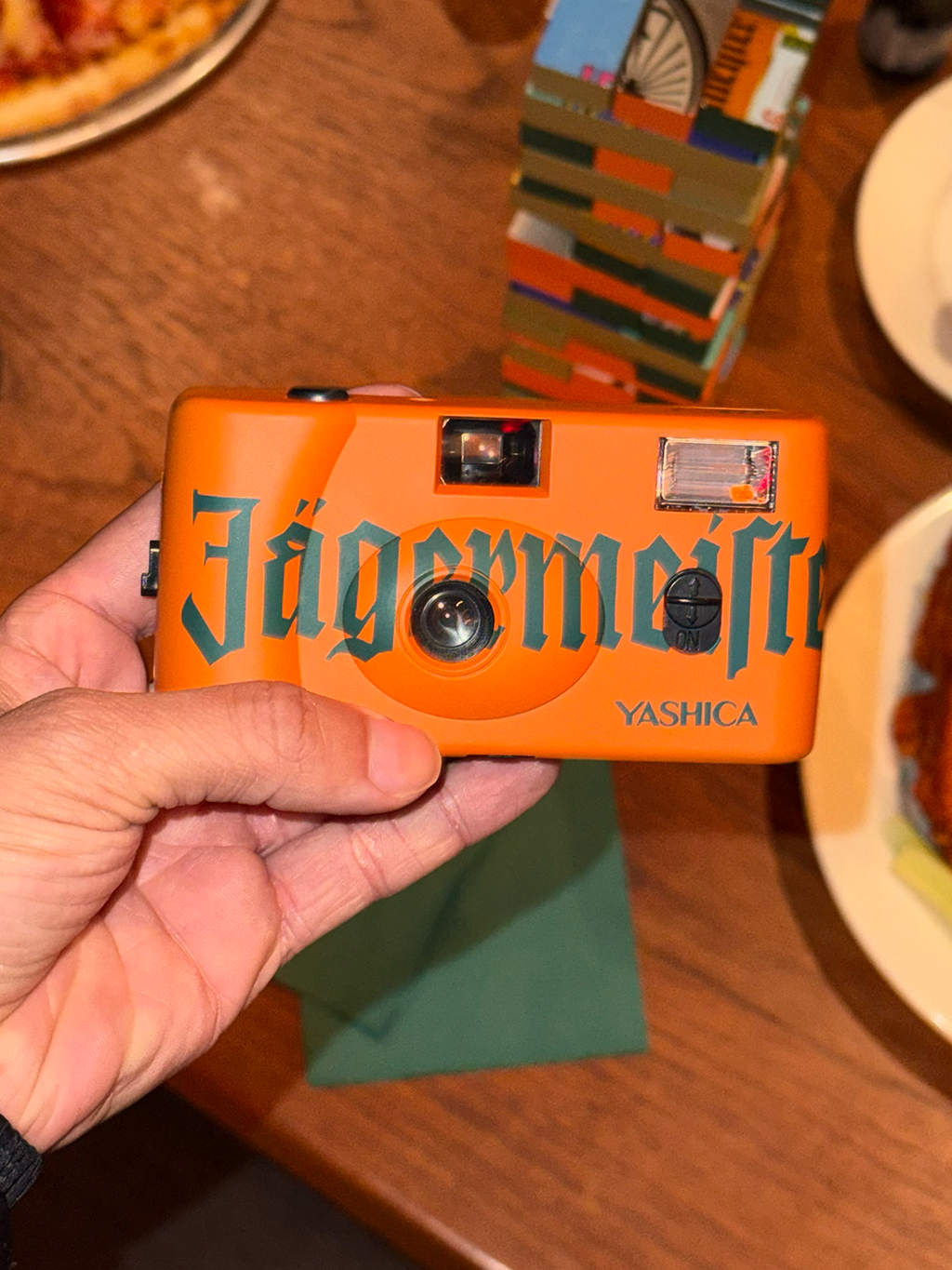 Narancssárga, „Jägermeister” márkajelzésű fényképezőgép, amelyet egy kéz tart egy faasztalon.